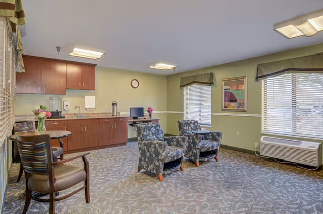 A well-lit common area with two floral patterned armchairs facing a small table, a round table with two chairs, and a kitchenette with wooden cabinets and a countertop. The room has light green walls, two windows with blinds and valances, a wall clock, a framed picture, and a carpeted floor.