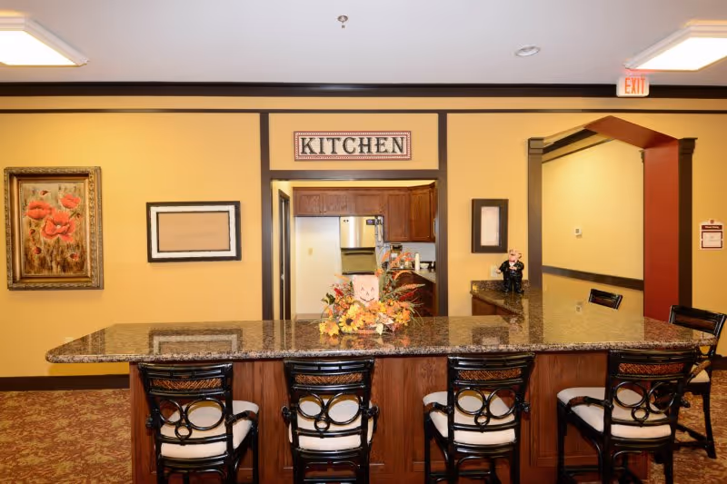 Interior view of a kitchen area in a retirement center with a granite countertop island and four cushioned chairs. The wall behind the island has a sign labeled 'KITCHEN' above an open doorway leading to the kitchen. There are decorative framed pictures on the walls and a floral centerpiece on the countertop. The room has warm yellow walls and patterned carpet flooring.