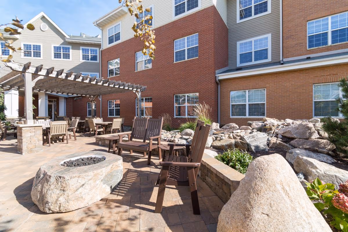 Outdoor patio area at SummerWood of Chanhassen featuring wooden chairs and tables under a pergola, a stone fire pit, and landscaping with rocks and plants in front of a multi-story brick and siding building.