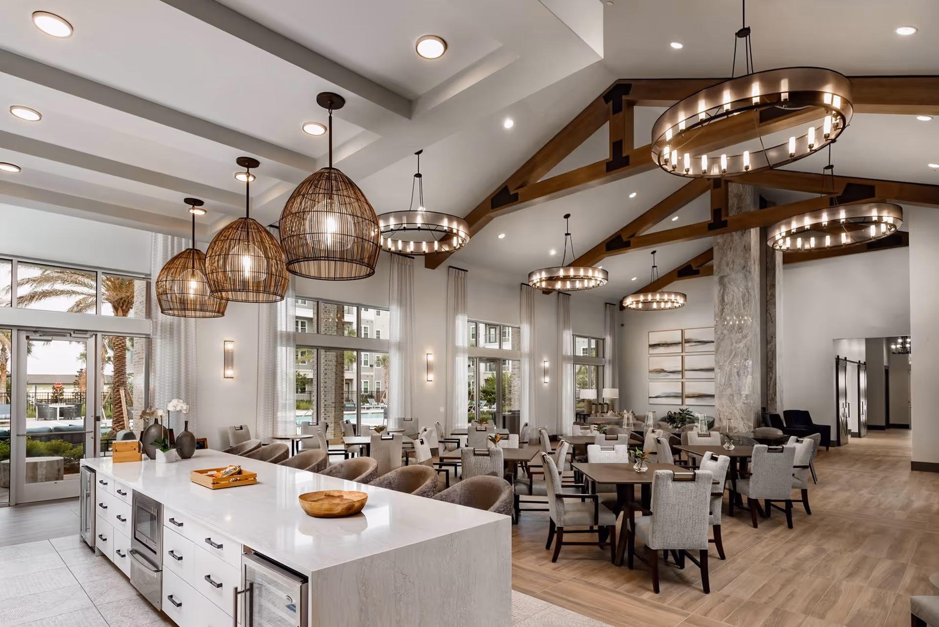 Spacious and modern dining area with multiple tables and chairs, large windows with sheer curtains allowing natural light, stylish pendant and chandelier lighting fixtures, and a long white countertop with bar stools and decorative items.