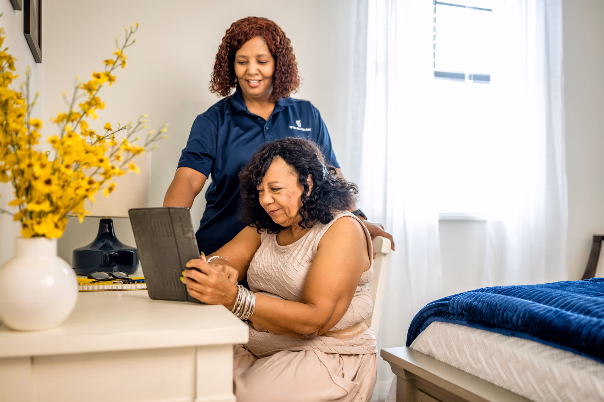 A caregiver in a navy blue Wellington Bay polo shirt stands behind a seated elderly woman who is using a tablet at a white desk in a bright bedroom. The room features a bed with a blue blanket, a white vase with yellow flowers, and a window with sheer white curtains.