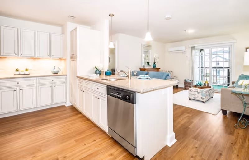 Bright and modern open-concept kitchen and living room area with white cabinetry, a kitchen island with a sink and dishwasher, wooden flooring, and a cozy living space with a sofa, ottoman, and sliding glass doors leading to a balcony.