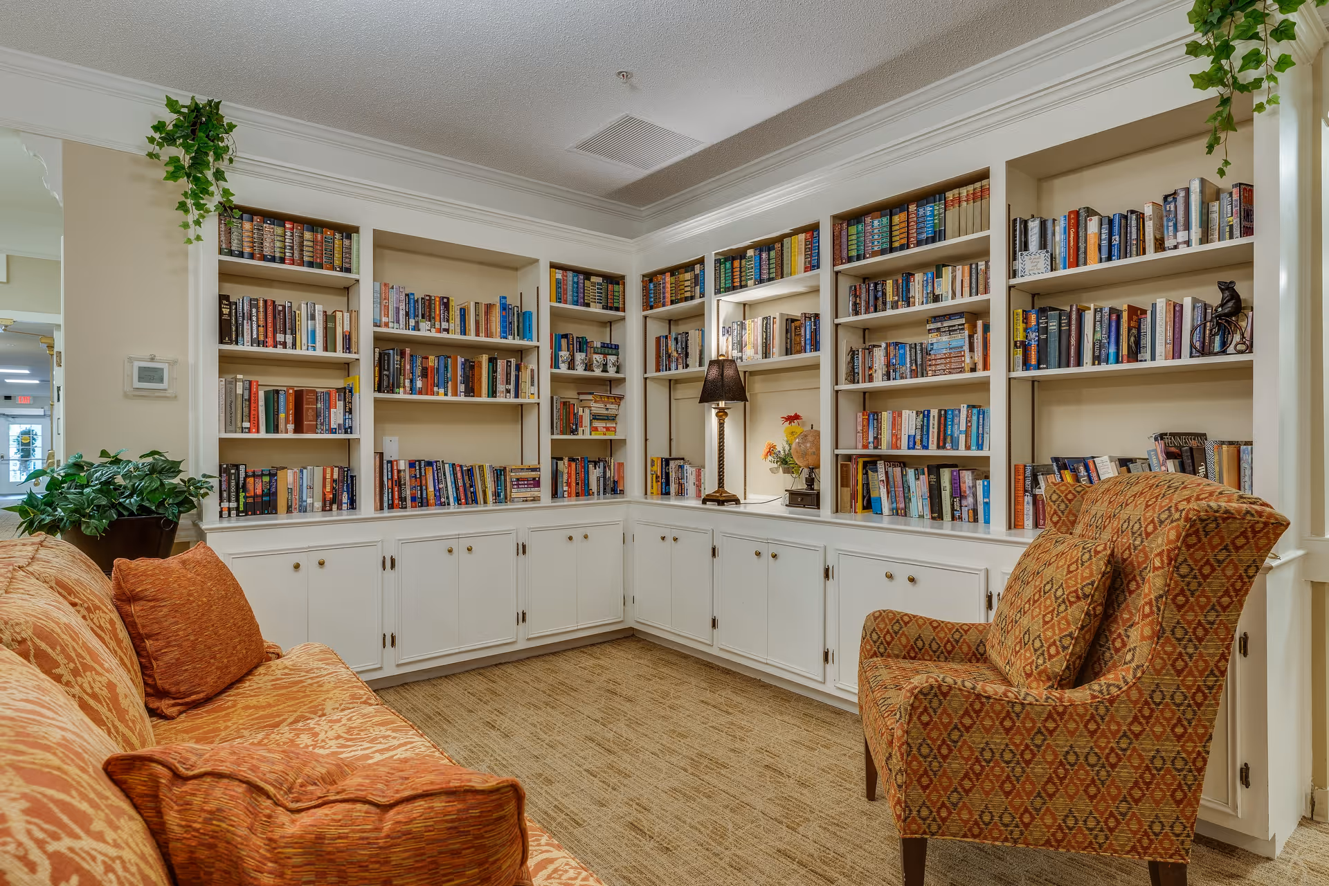 Cozy reading nook in a senior living facility featuring built-in white bookshelves filled with books, a patterned orange armchair, an orange sofa with matching pillows, a table lamp, and green plants hanging and placed on the shelves.