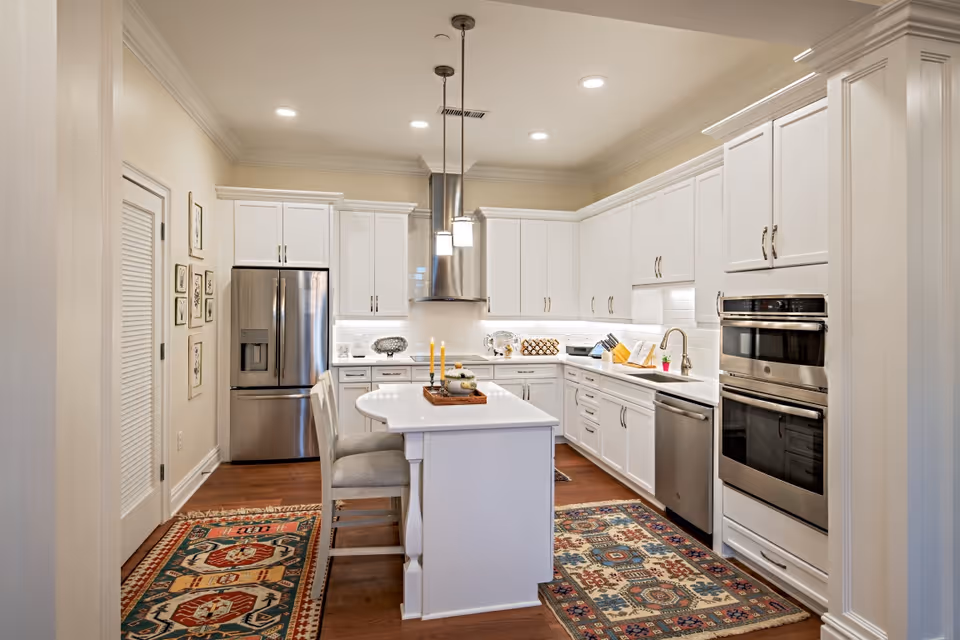 Bright modern white kitchen with a center island, stainless steel appliances, pendant lights, and patterned rugs.