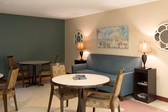 A cozy seating area with a blue sofa against a beige wall, flanked by two dark wooden side tables each with a lamp. Above the sofa is a colorful abstract painting. In front of the sofa are round tables with patterned chairs arranged around them. The room has a green accent wall and a light-colored floor.