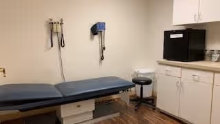 A medical examination room with a padded examination table, wall-mounted blood pressure cuff and otoscope, a small stool, a sink, and white cabinets with a black microwave on the countertop.