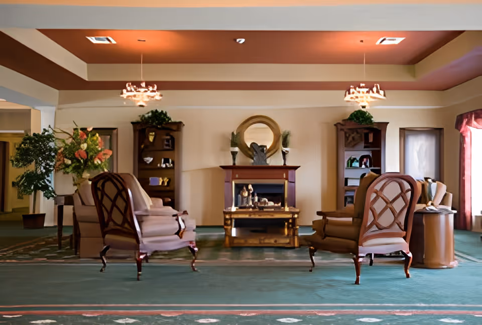 A spacious living room area with four upholstered armchairs arranged around a wooden coffee table and a fireplace. The room features two wooden bookshelves with decorative items, a round mirror above the fireplace, potted plants, and two hanging light fixtures from a recessed ceiling. Large windows with curtains allow natural light into the room.