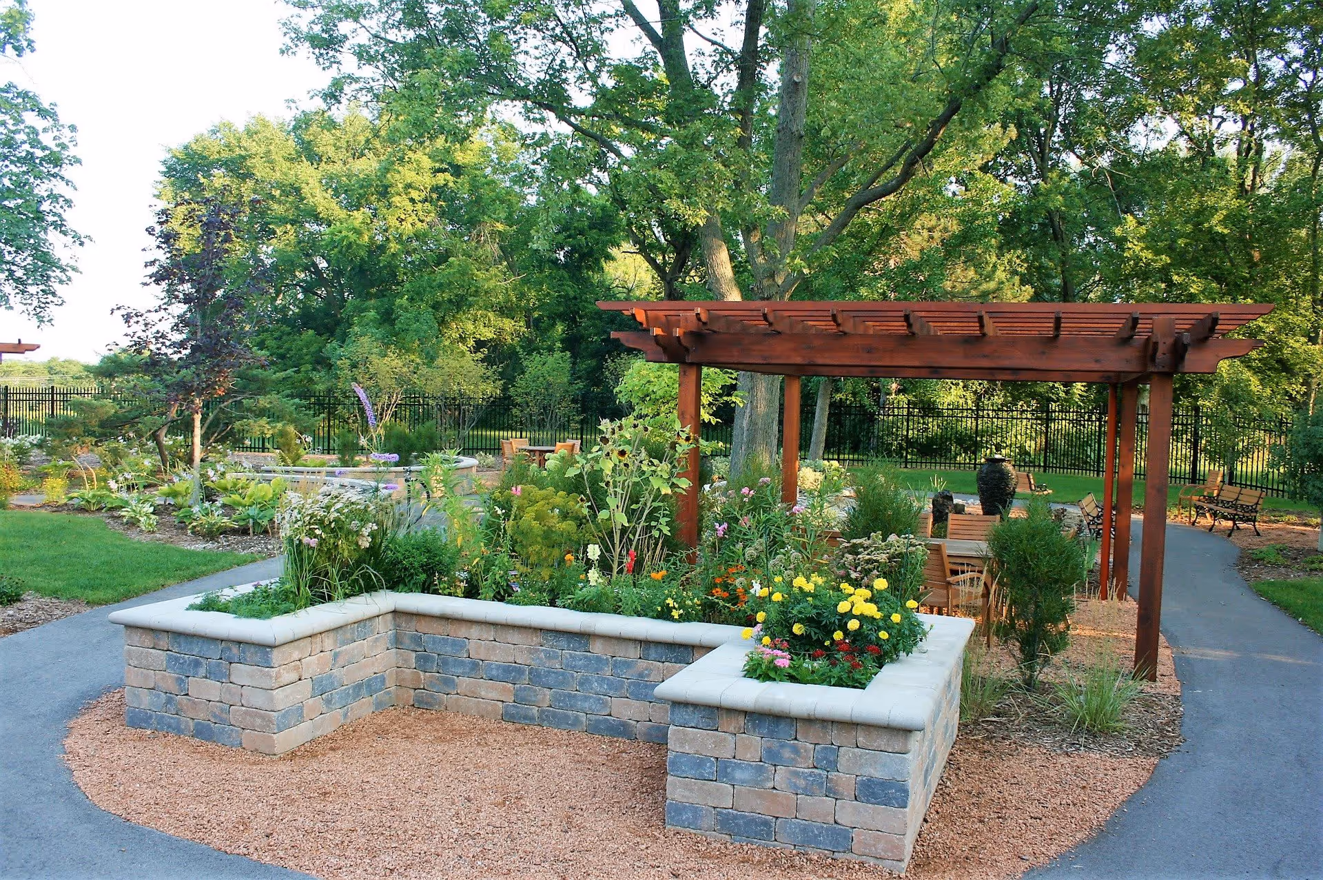 Outdoor garden area with a stone raised flower bed filled with various colorful flowers and plants. Behind the flower bed is a wooden pergola with seating and tables underneath. The area is surrounded by trees and greenery with a paved pathway leading through the garden.
