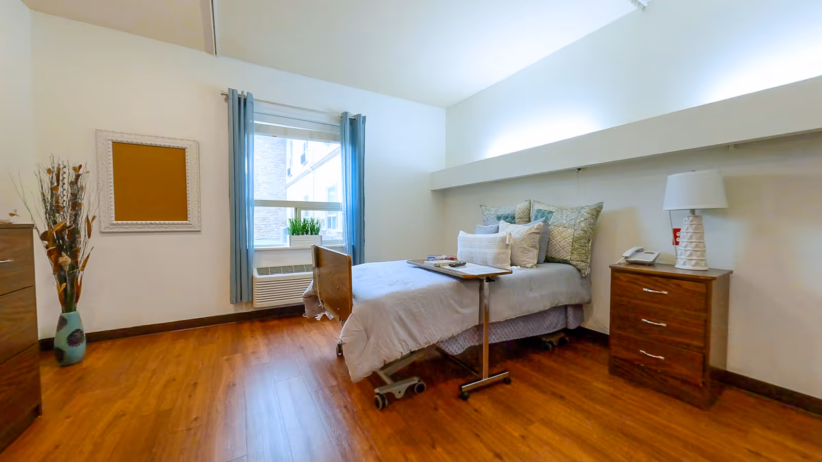 A bright and clean bedroom in a senior living facility with a single bed on wheels, a bedside table with a lamp and telephone, a window with blue curtains, a wooden dresser, a vase with decorative branches, and a corkboard on the wall.