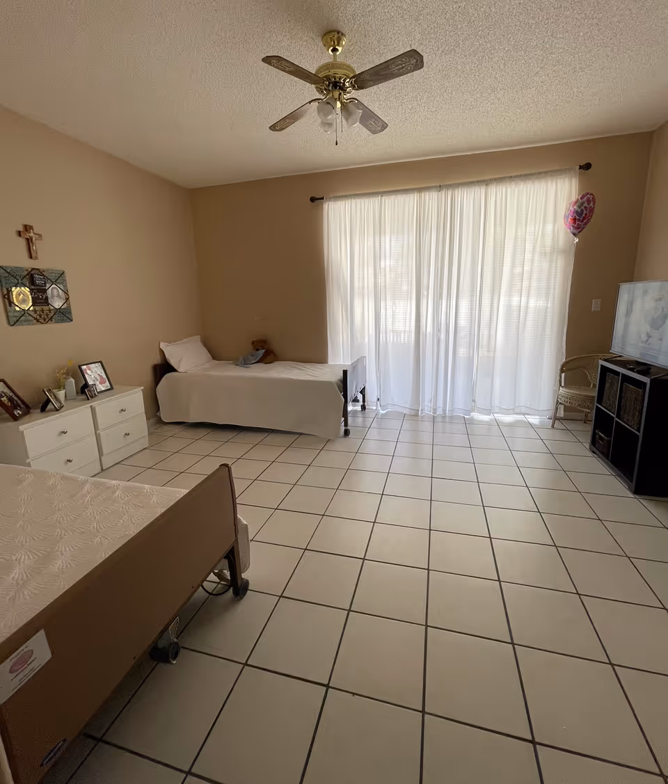 A simple bedroom with two single beds, one against the left wall and one in the foreground. The room has beige walls, a white tile floor, a ceiling fan with lights, and a large window covered with sheer white curtains. There is a white dresser with framed photos and decorations on the left side, a small TV on a black stand on the right, and a wicker chair next to it. A pink heart-shaped balloon is tied near the window.