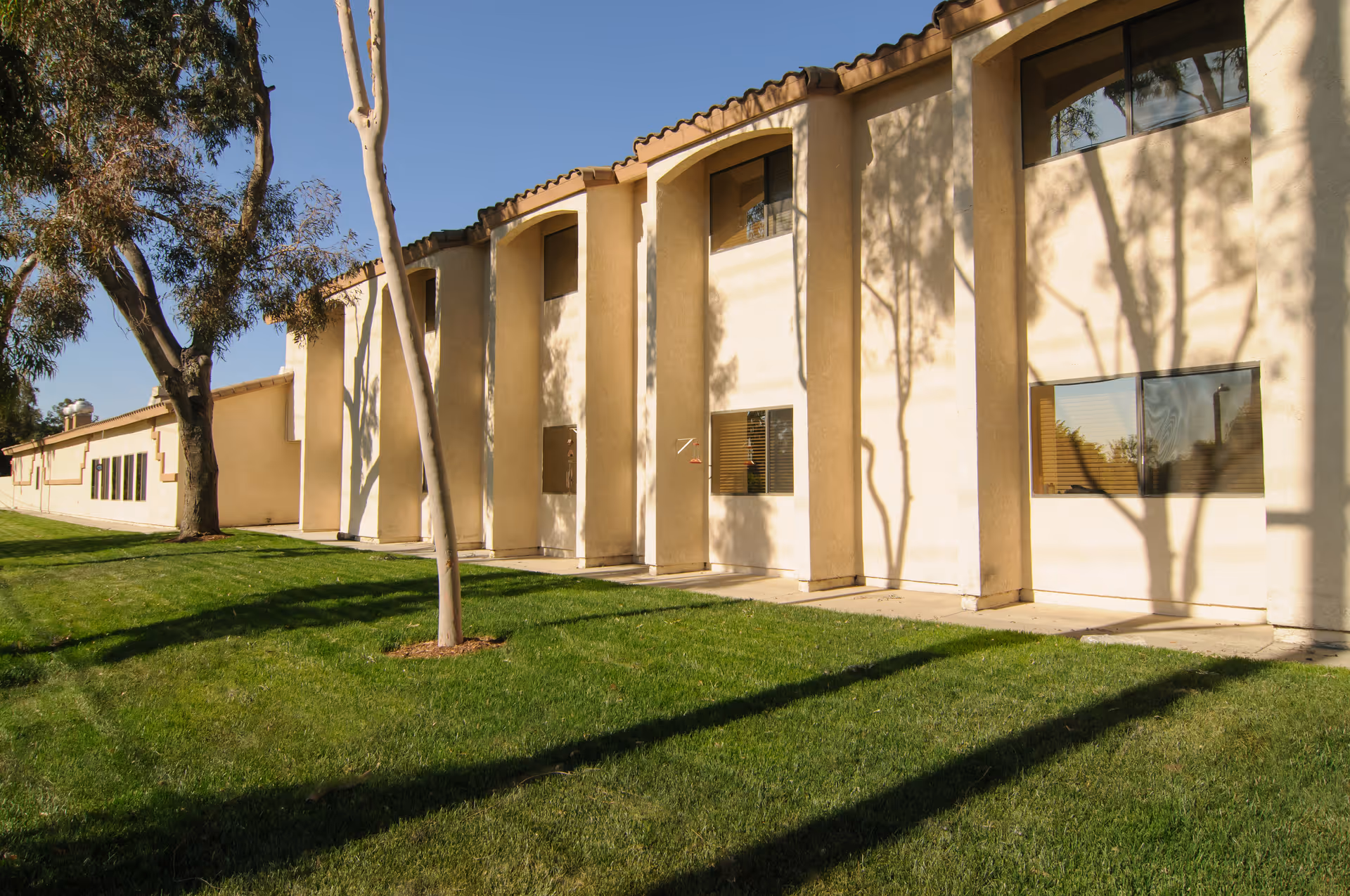 Exterior view of a beige two-story building with arched architectural features, several windows, and a well-maintained green lawn with trees casting shadows on the building and grass under a clear blue sky.