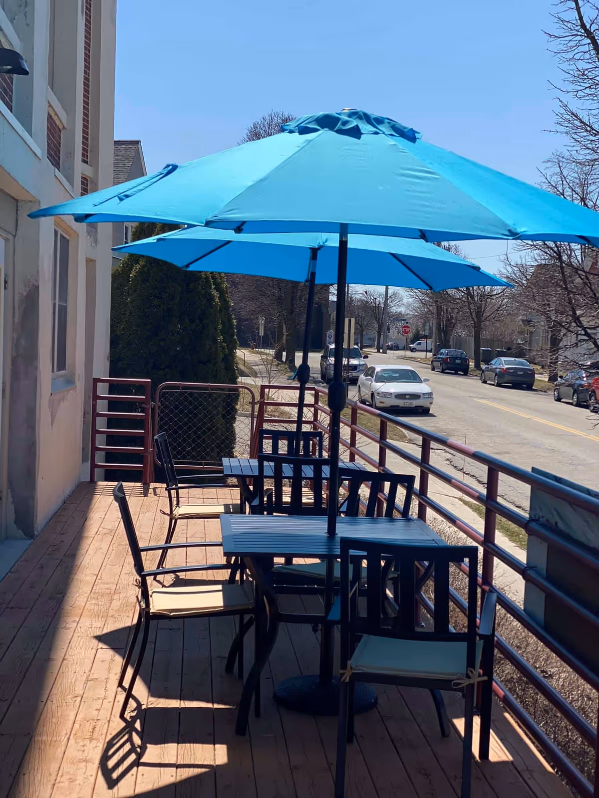 Outdoor patio area with wooden deck featuring black metal tables and chairs with beige cushions. Two large blue umbrellas provide shade. The patio is adjacent to a building and overlooks a street with parked cars and leafless trees under a clear blue sky.