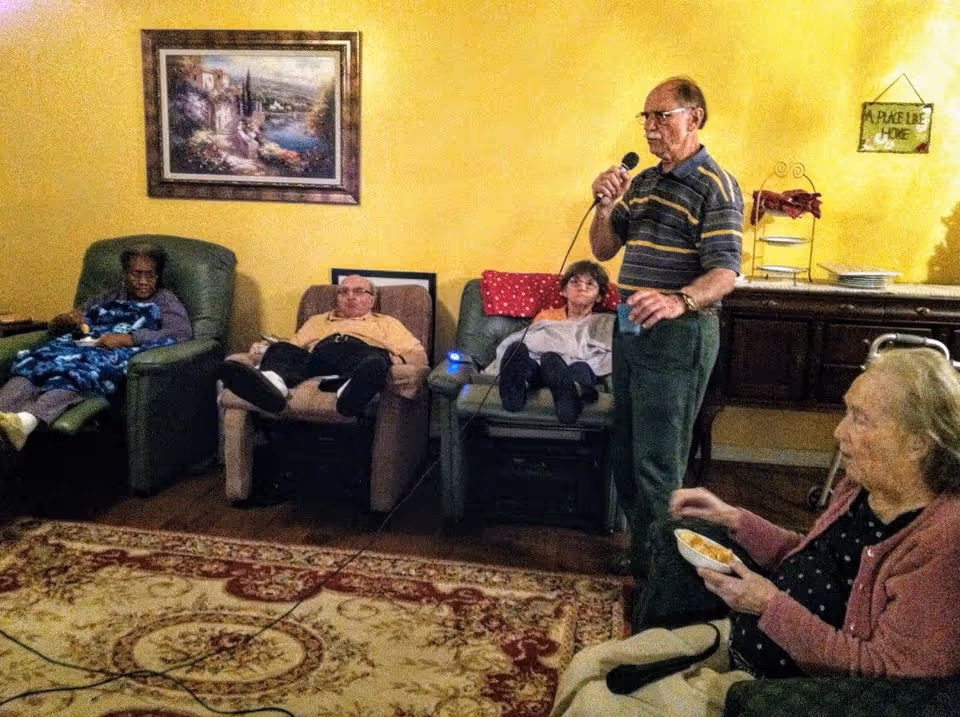 A group of elderly people in a living room setting. Three individuals are seated in recliners against a yellow wall, one man is standing and holding a microphone, and another woman is seated in the foreground eating from a bowl. The room has a patterned rug on the floor, a framed painting on the wall, and a cabinet with plates and a decorative sign that reads 'A Place Like Home'.