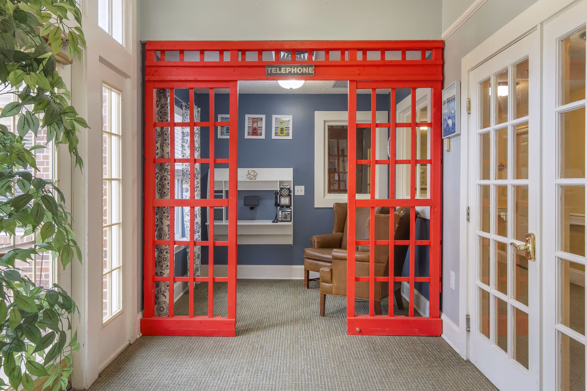 Interior view of a room in Brookdale Burlington featuring a red British-style telephone booth frame without a door. Inside the booth area, there is a small white desk with a vintage black rotary phone mounted on the wall. A brown leather armchair is positioned to the right of the desk. The walls are painted blue with framed pictures hanging above the desk. To the left, there is a large window with patterned curtains and a green leafy plant near the window. To the right, there is a white door with glass panes.