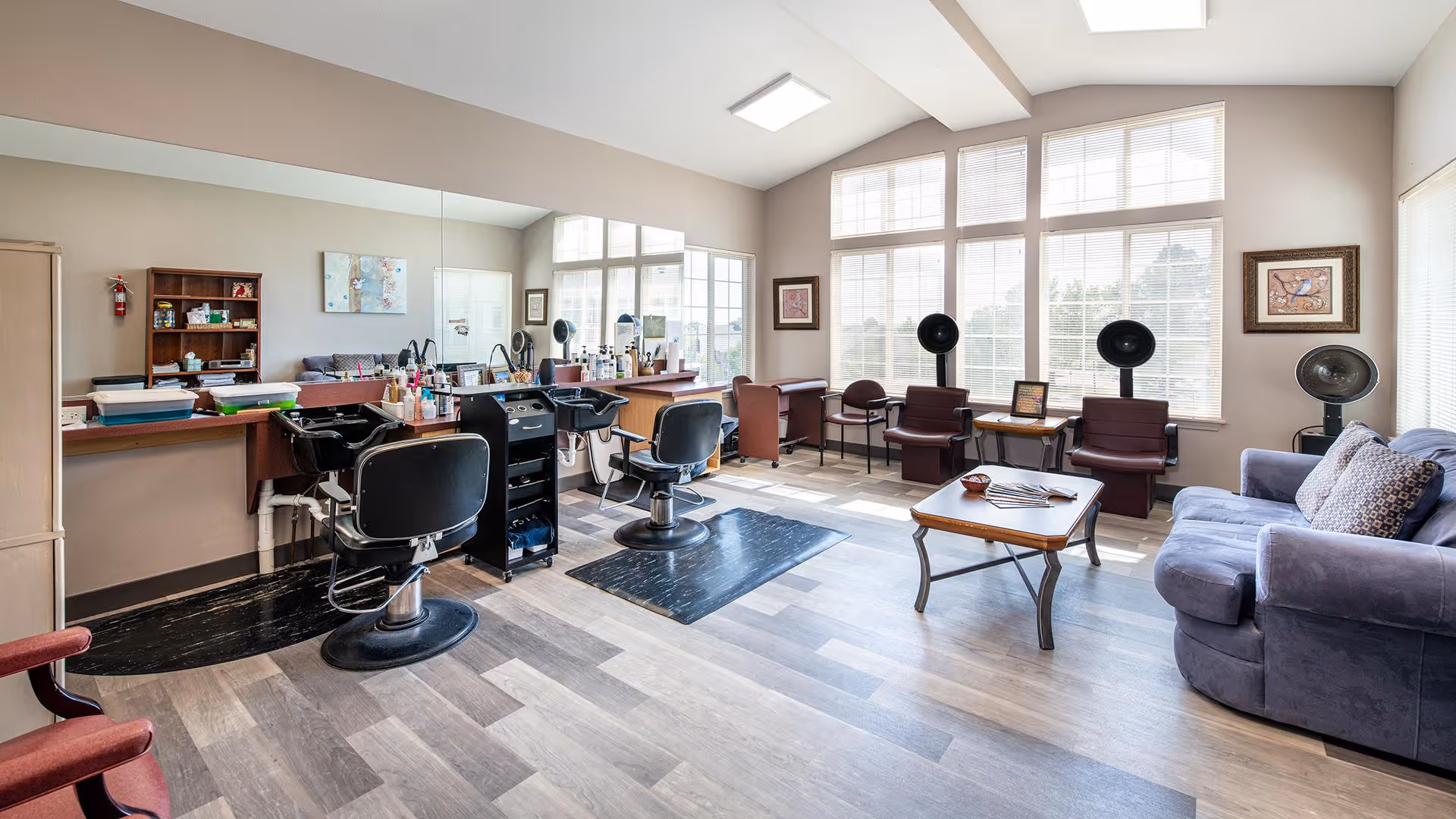 A bright and spacious salon area with large windows letting in natural light. The room features two black salon chairs in front of mirrors and sinks for hair washing, a small black rolling cart with hair products, and a wooden coffee table with magazines. There are also several salon hair dryers and comfortable seating including a blue couch and multiple chairs. The walls are painted a soft beige, and the floor has a light wood pattern.