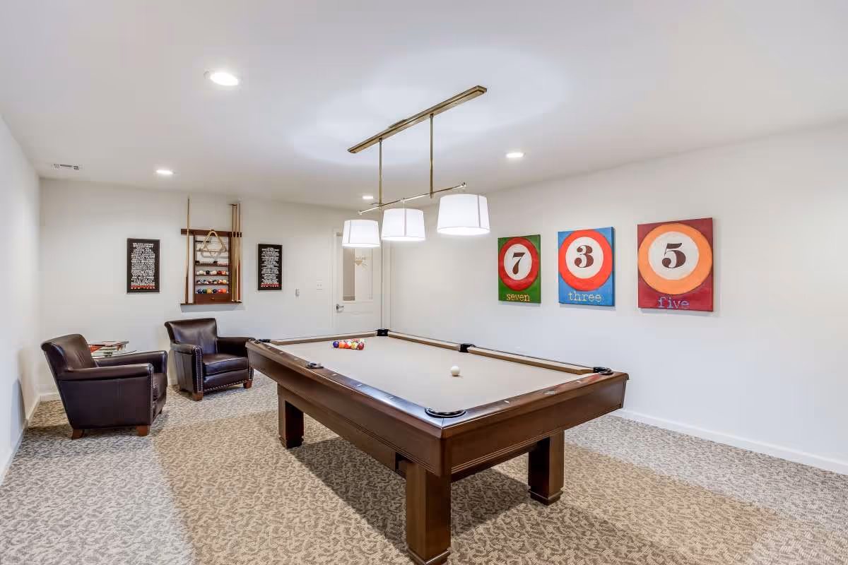 A well-lit game room featuring a pool table with balls arranged for a game. The room has patterned carpet flooring, two dark leather armchairs with a small side table between them, and a wall-mounted rack holding pool cues and additional balls. Three colorful paintings of billiard balls numbered seven, three, and five hang on the wall. A modern light fixture with four lamps hangs above the pool table.