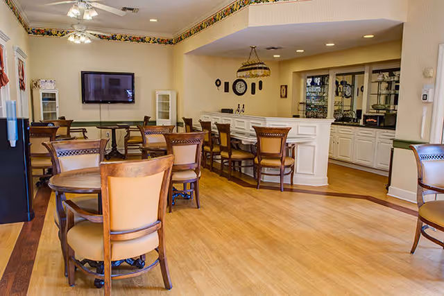 A spacious dining area with several wooden tables and chairs arranged on a light wood floor. The room features a mounted flat-screen TV on the far wall, a white counter with additional chairs, and decorative wall accents including a clock and hanging plates. The ceiling has recessed lighting and ceiling fans, and there is a glass-front cabinet behind the counter.