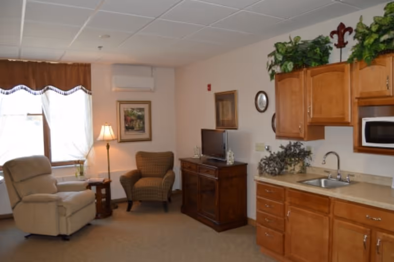Interior common room with a recliner and armchair beside a small TV cabinet and a kitchenette with wooden cabinets and a sink.