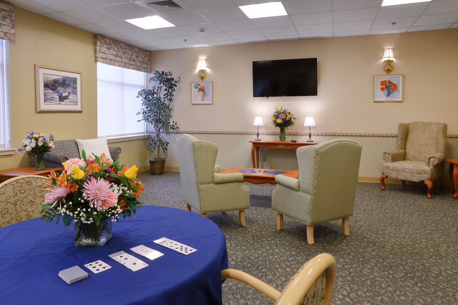 A cozy living room area in a senior living facility with a blue tablecloth-covered table in the foreground holding a vase of colorful flowers and playing cards. The room features two green upholstered armchairs facing a wall-mounted flat-screen TV, a wooden coffee table, a beige armchair, framed artwork on the walls, a potted plant near the window, and soft lighting from wall sconces and table lamps.