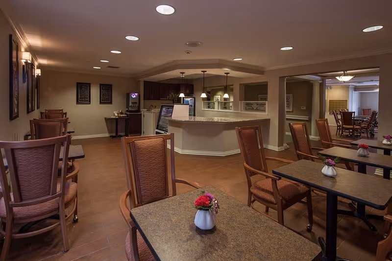 Interior view of a dining area in Waltonwood Cherry Hill featuring several tables with brown chairs, small flower vases on the tables, warm lighting, and a counter area with pendant lights in the background.