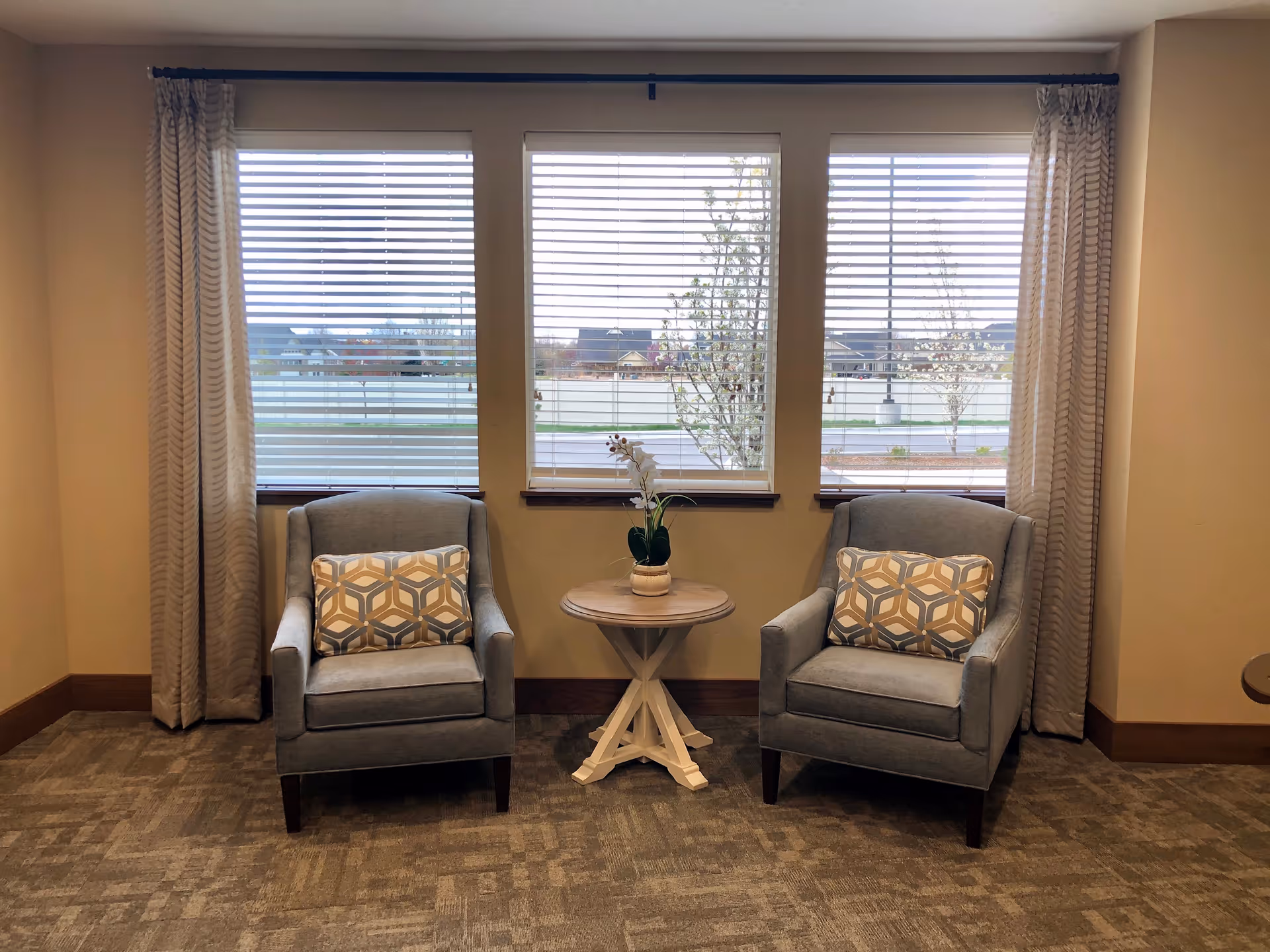 Two upholstered armchairs with patterned pillows flank a small round table with a plant in front of three large windows with blinds and curtains.