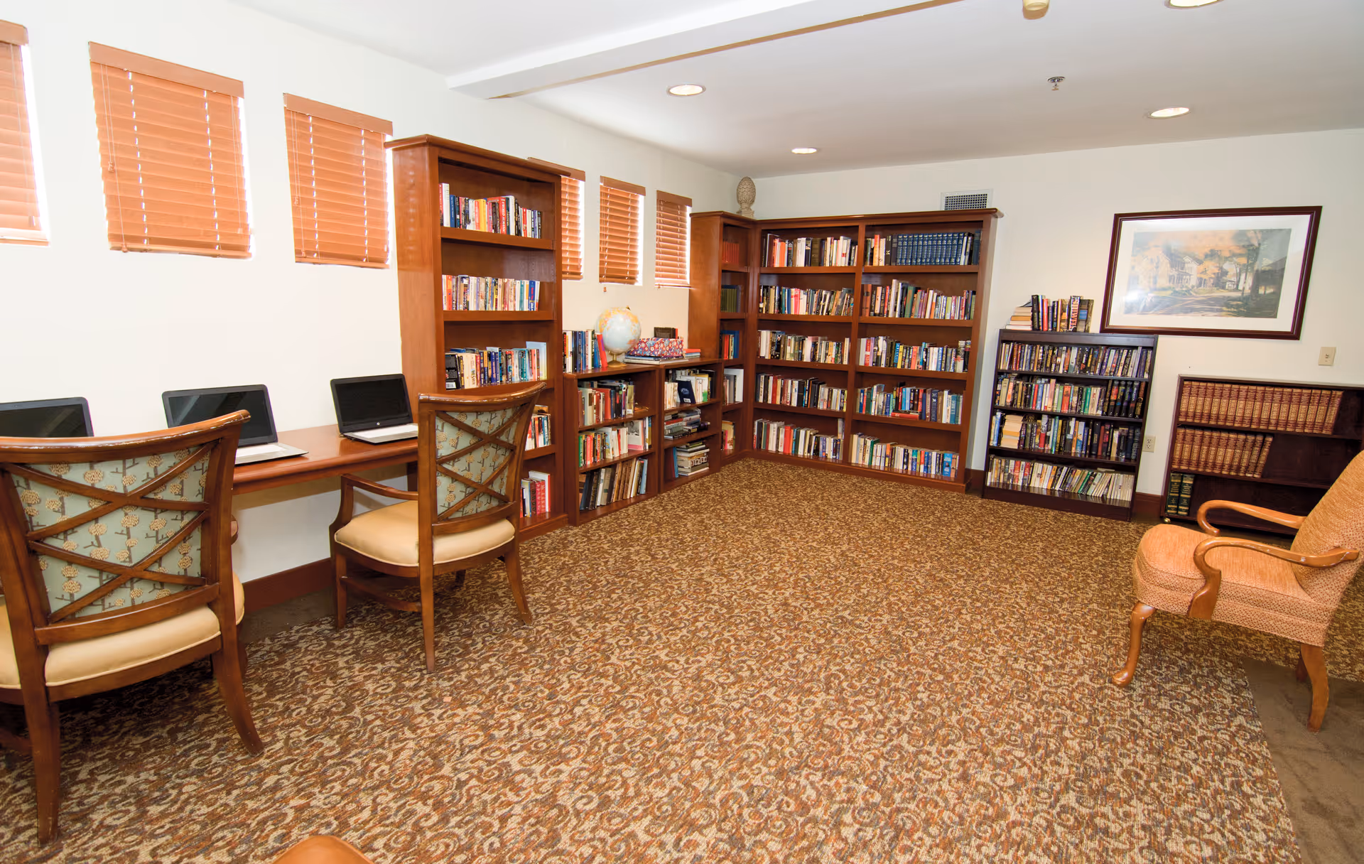 A cozy library or reading room with multiple wooden bookshelves filled with books, a few laptops on a long wooden desk with chairs, a globe, and a framed picture on the wall. The room has patterned carpet and small windows with blinds.