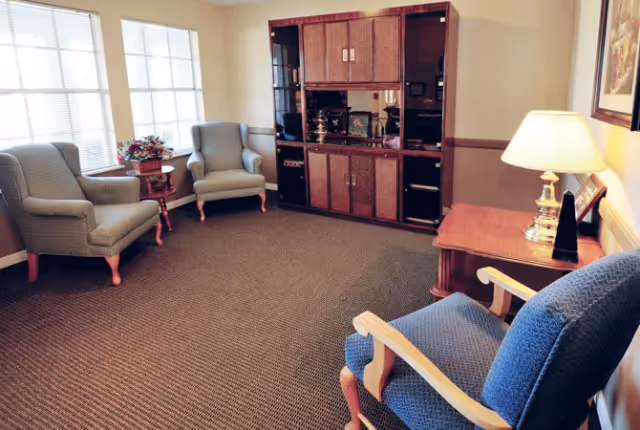 A cozy living room area with three upholstered armchairs arranged around a wooden entertainment cabinet. A side table with a lamp and framed picture is next to one chair, and another small table with a flower arrangement is placed between two chairs near large windows letting in natural light.