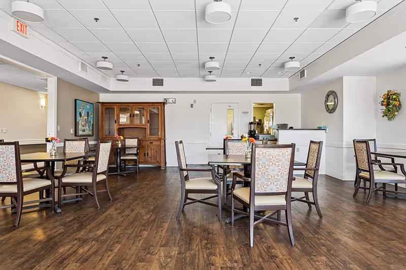 A dining room with multiple wooden tables and chairs with patterned upholstery. The room has wood flooring, a white ceiling with round light fixtures, and a wooden cabinet against the far wall. There is a clock on the right wall and a floral wreath decoration. A doorway leads to another room with kitchen equipment visible.