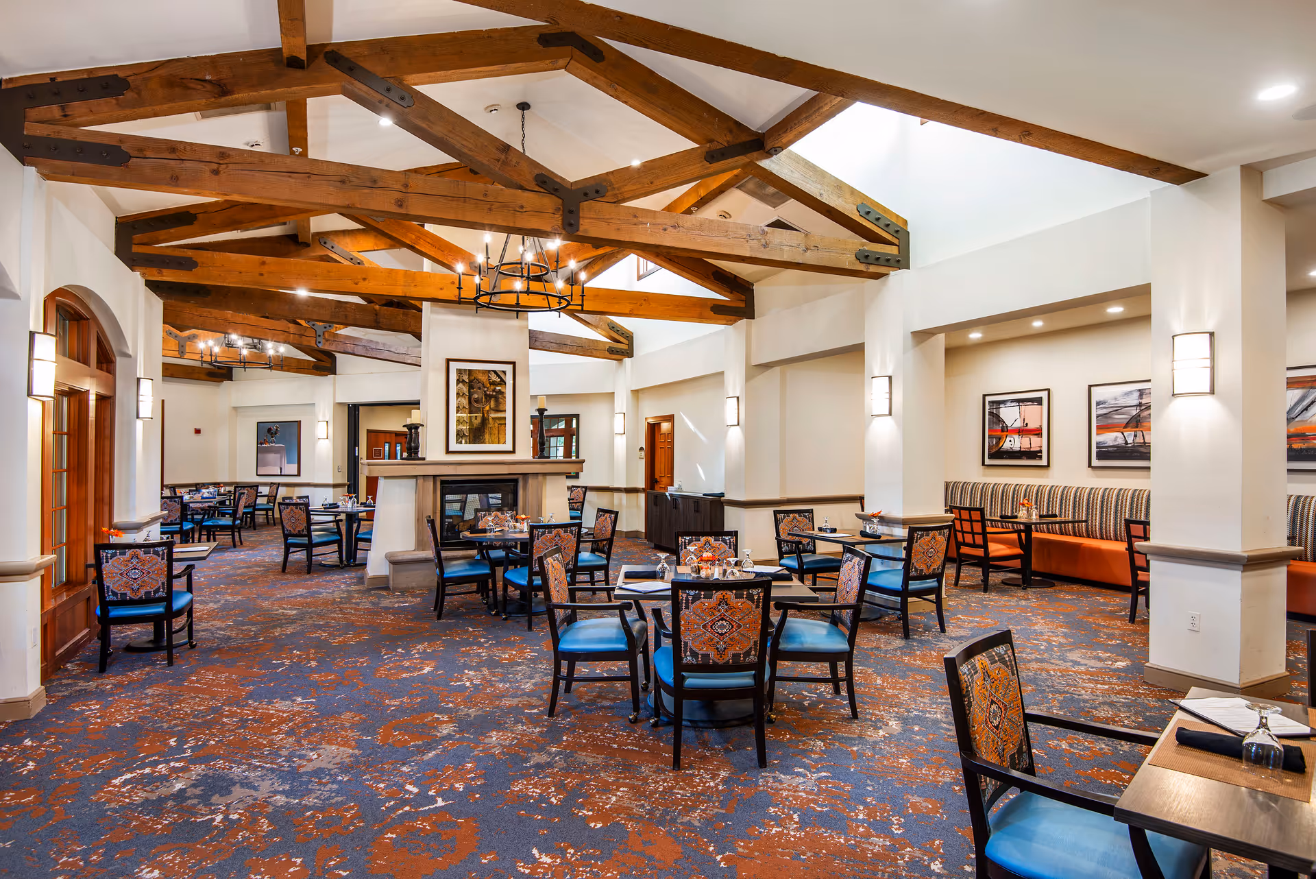 Spacious dining room with exposed wooden ceiling beams, tables and chairs arranged around a central fireplace.