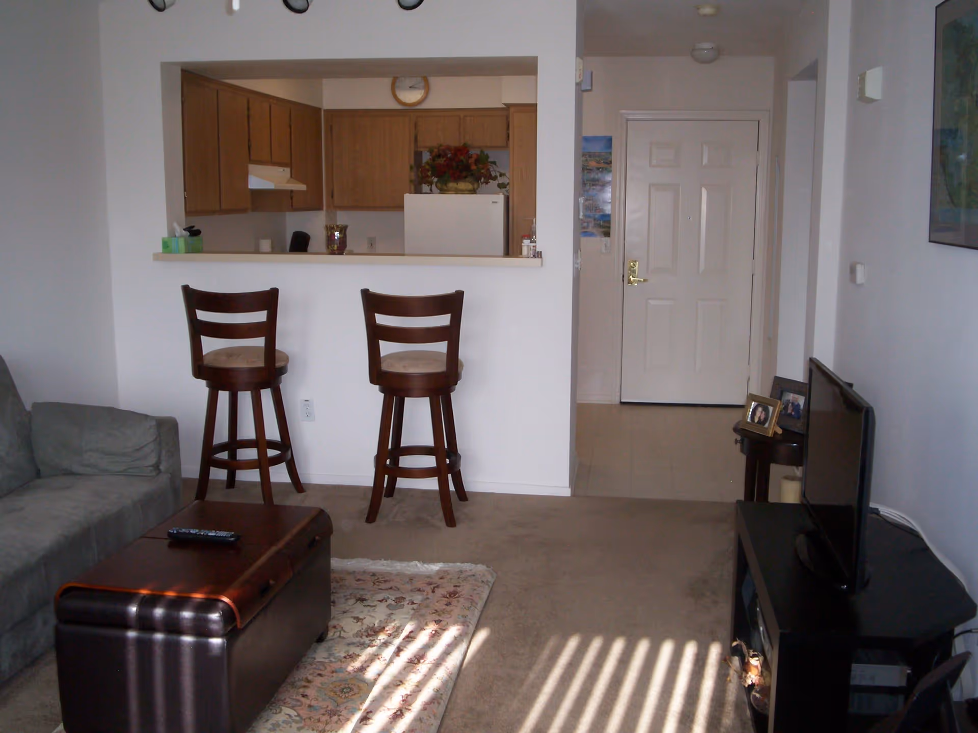 Living room showing two bar stools at a kitchen pass-through, a couch, ottoman, and TV with the entry door in view.
