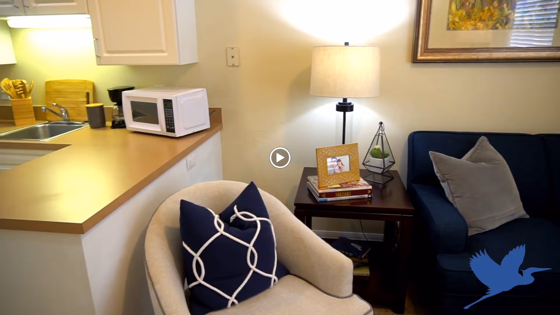 Interior view of a cozy living space featuring a beige armchair with a navy blue pillow, a wooden side table with a lamp, books, a framed photo, and a decorative plant. To the left is a kitchen counter with a microwave, coffee maker, and utensils near a sink.