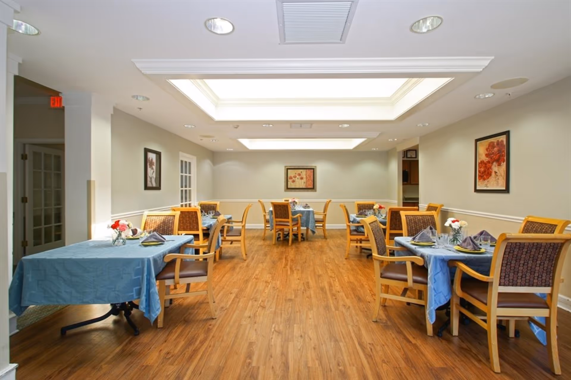 A bright dining room with multiple tables covered in blue tablecloths and wooden chairs beneath recessed skylights.
