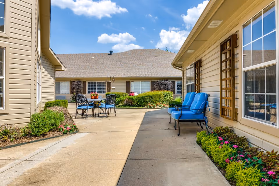 Outdoor patio area at a senior living facility with a concrete walkway, blue cushioned chairs along the building wall, a round table with chairs and a flower pot, surrounded by bushes and flowers under a partly cloudy sky.
