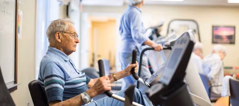 An elderly man using a stationary exercise bike in a fitness room, with other seniors exercising on treadmills and seated in the background.