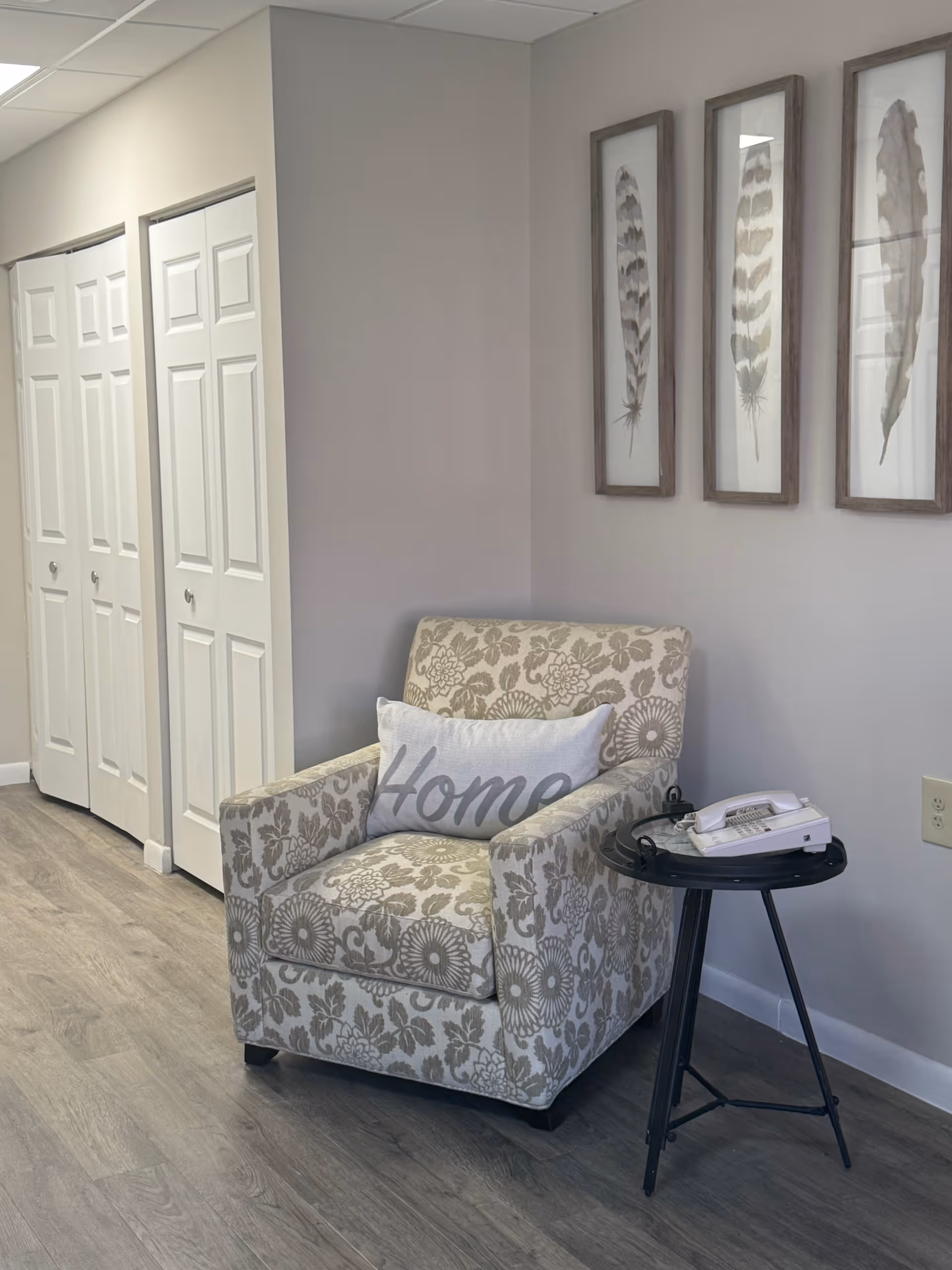 A cozy corner with a patterned armchair featuring a pillow that says 'Home', next to a small black side table holding a white landline telephone. The wall behind has three framed feather artworks, and there are white double closet doors along the adjacent wall. The floor is wood with a light gray finish.