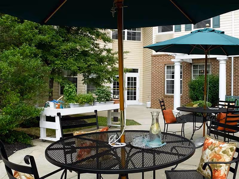 Outdoor patio area with black metal mesh tables and chairs, some with floral cushions. Green umbrellas provide shade over the tables. In the background, there is a white planter box with gardening tools and plants, along with a building featuring brick and beige siding with white columns and windows.