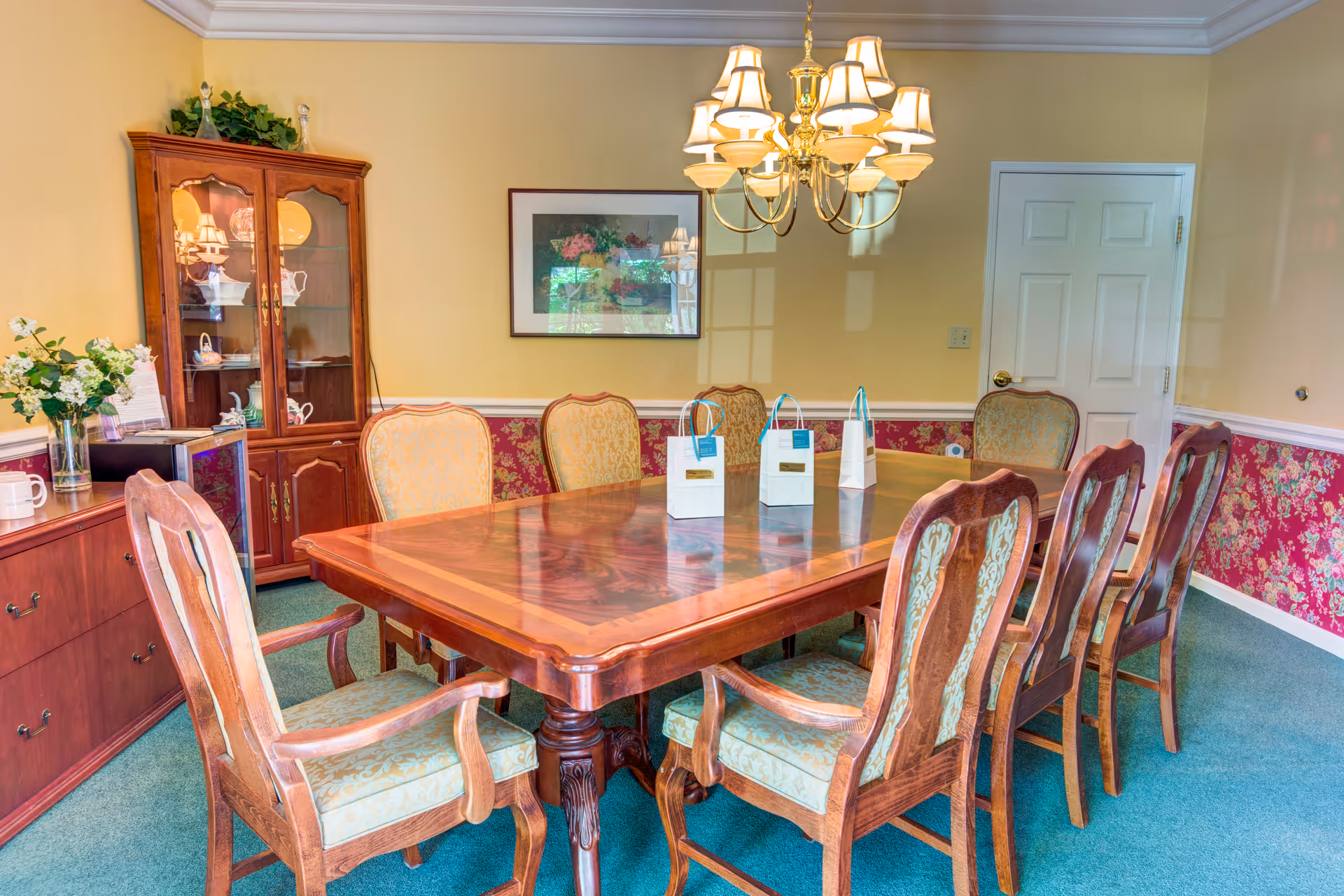 A traditional dining room with a large wooden dining table surrounded by eight upholstered chairs. The room features a chandelier hanging above the table, a wooden cabinet with glass doors displaying dishes, a sideboard with a vase of flowers, and floral wallpaper on the lower half of the walls. A framed picture hangs on the wall, and a closed white door is visible in the background.