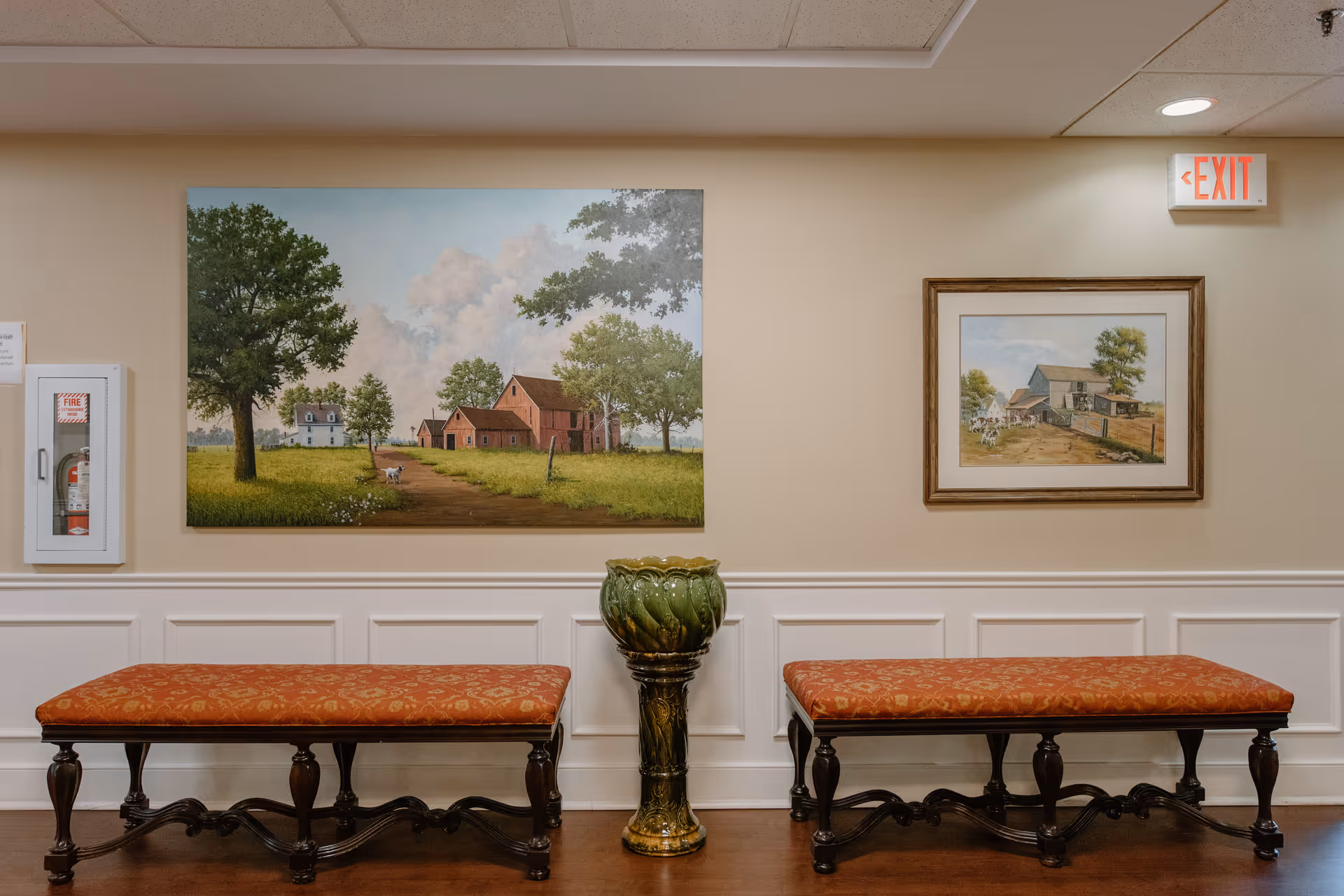 Interior hallway with two red cushioned benches with dark wooden legs placed against a beige wall with white wainscoting. Between the benches is a decorative green ceramic pedestal. Above the benches are two framed paintings depicting rural farm scenes with trees, barns, and houses. A fire extinguisher cabinet is mounted on the left wall, and an illuminated exit sign is visible on the upper right.