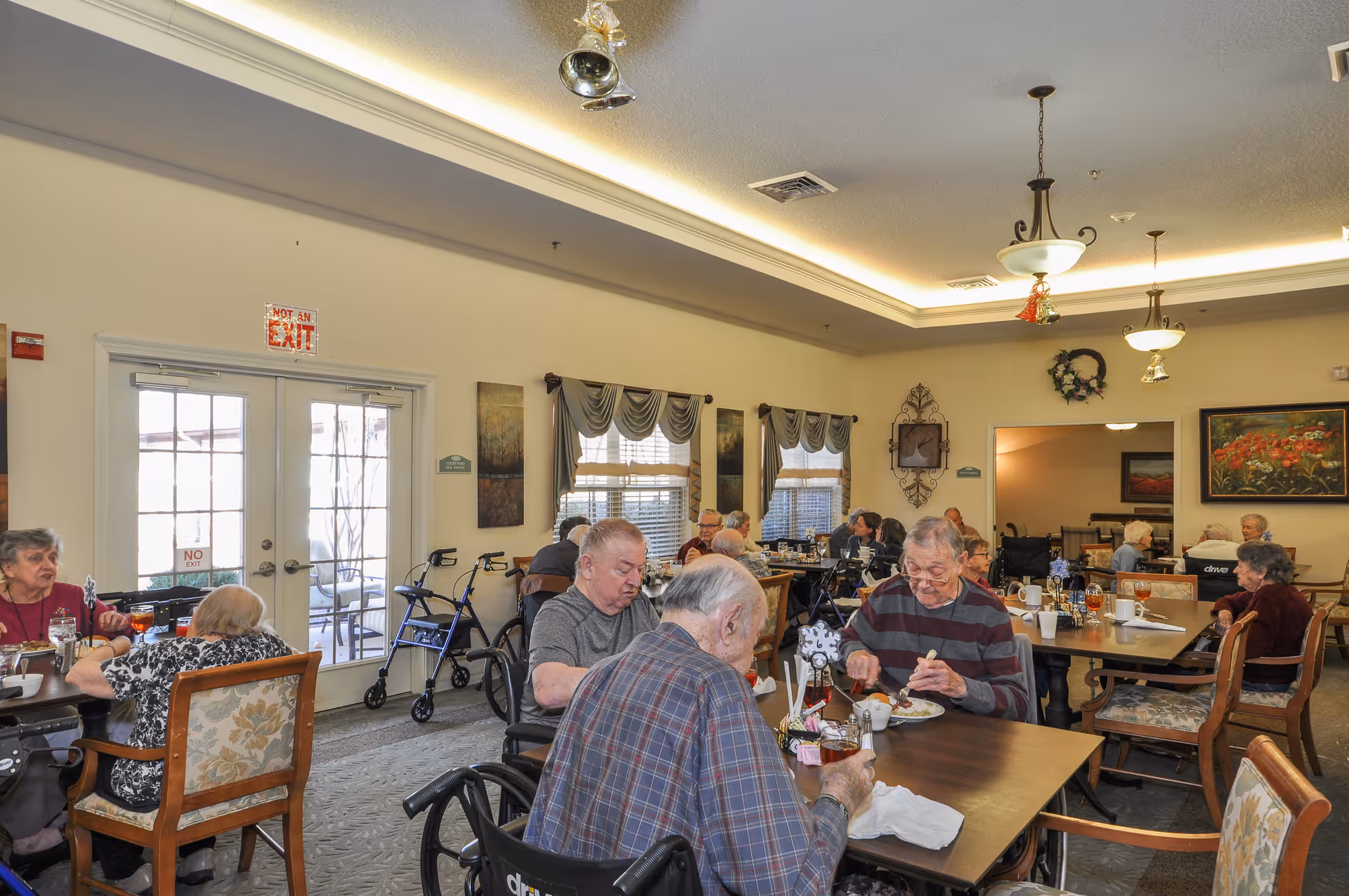 A dining room in a senior living facility with elderly residents seated at tables eating and socializing. The room has large windows with curtains, framed artwork on the walls, and ceiling lights. Walkers and wheelchairs are visible near some residents.