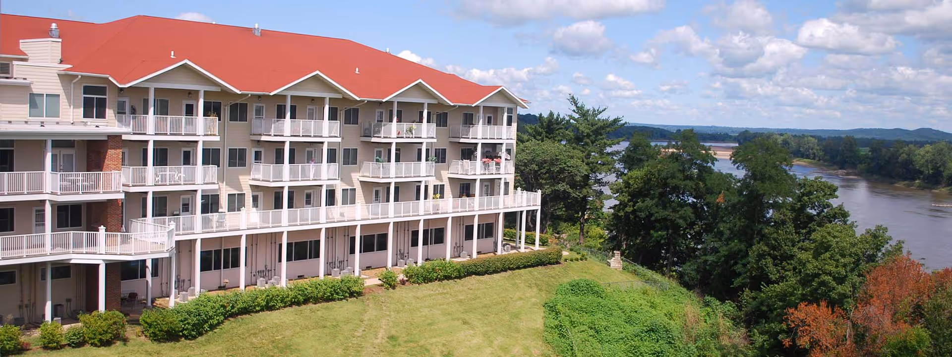 Multi-story senior living building with balconies overlooking a lawn and a river under a partly cloudy sky.