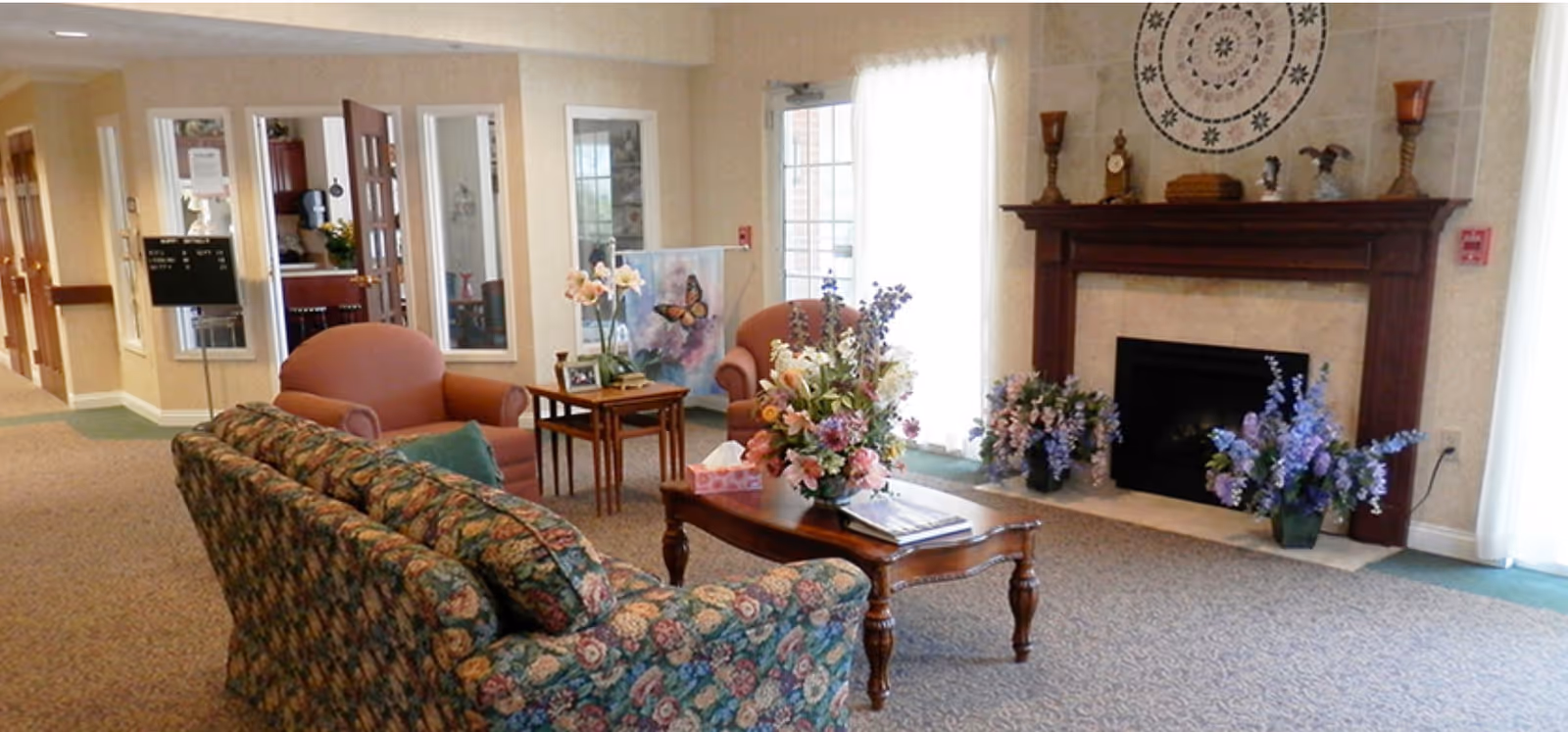 Carpeted senior living room lounge with a floral sofa and armchairs around a coffee table topped with flower arrangements in front of a fireplace.