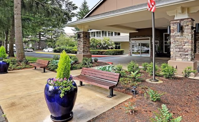 Outdoor view of the entrance area to Windsor Square Senior Living facility featuring a covered drop-off zone supported by stone pillars, landscaped garden beds with small shrubs, two wooden benches on a concrete walkway, a large blue planter with flowers and greenery, and an American flag on a flagpole.