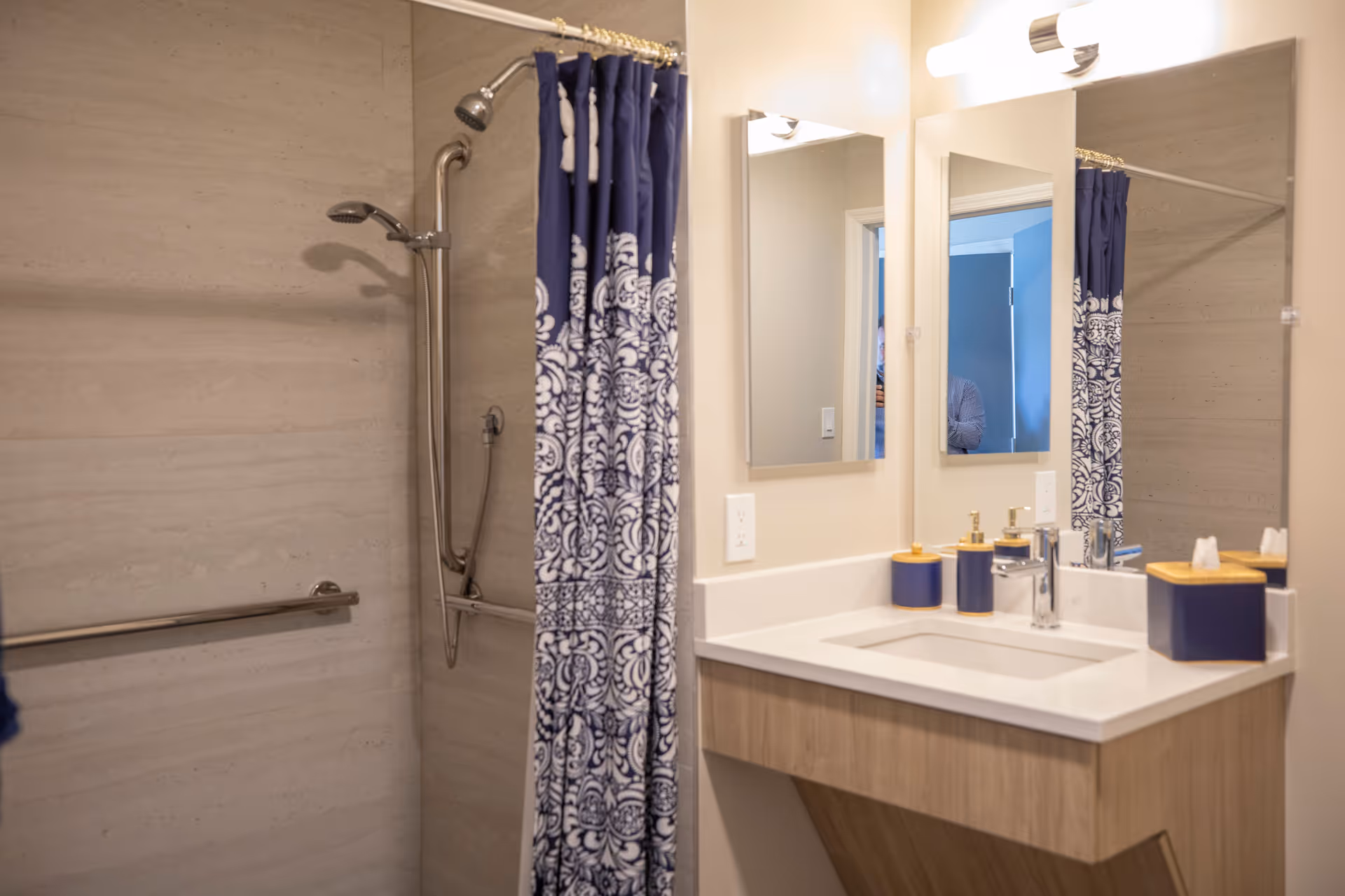A bathroom with a shower area featuring a blue and white patterned shower curtain, a handheld showerhead, and grab bars. Next to the shower is a vanity with a white countertop, a sink, and three blue and gold bathroom accessories. Above the vanity is a large mirror with a light fixture.