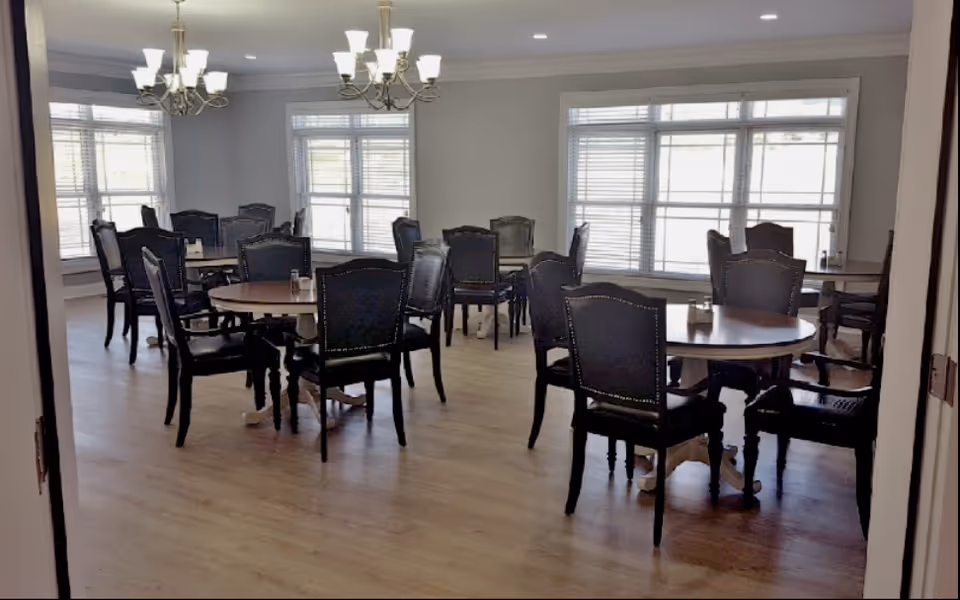 Dining room with several round tables and upholstered chairs beneath chandeliers and large windows.