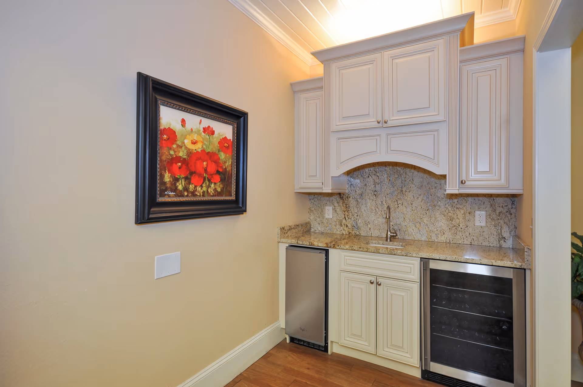 A small kitchenette area with cream-colored cabinets, a granite countertop and backsplash, a small sink with a faucet, a mini refrigerator, and a wine cooler. On the left wall, there is a framed painting of red and yellow flowers. The floor is wooden and the ceiling has recessed lighting.