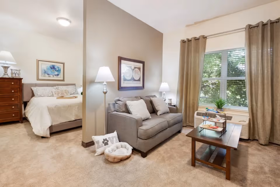 A cozy living space in a senior living facility featuring a gray sofa with white pillows, a wooden coffee table with snacks, a floor lamp, and a window with beige curtains. To the left, there is a bedroom area with a bed, a wooden dresser, a table lamp, and framed artwork on the walls. The room has beige carpeting and neutral-colored walls.