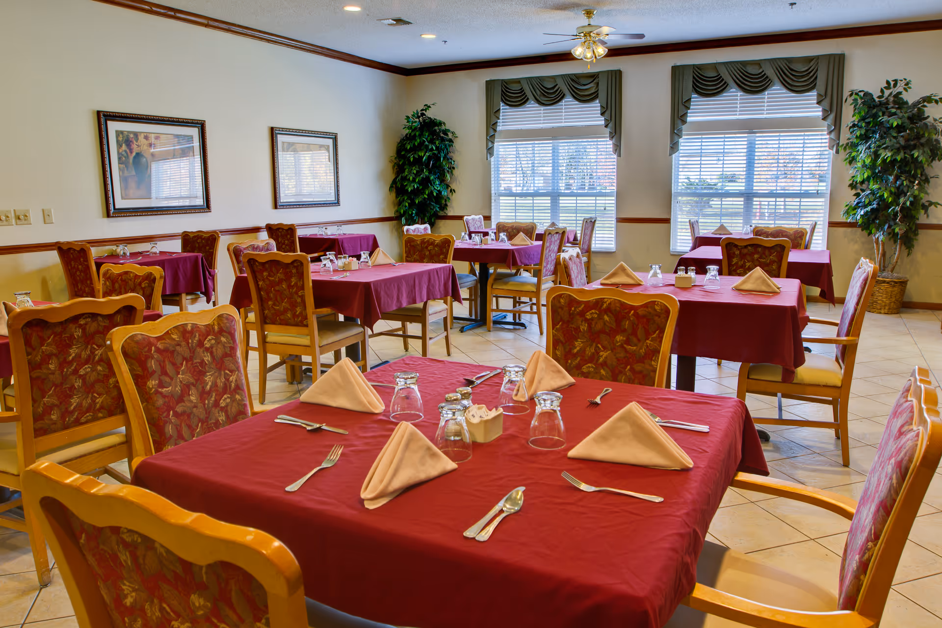 Dining room with multiple tables covered in red tablecloths, place settings with folded napkins and upholstered chairs near windows.