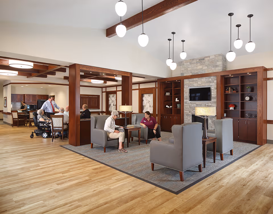 A spacious and well-lit assisted living facility common area with wooden beams and flooring. The room features a seating area with four gray armchairs around a small table, two lamps, and built-in wooden shelves with a stone fireplace and a mounted TV. In the background, several elderly residents and staff members are interacting near a dining area and kitchen space.