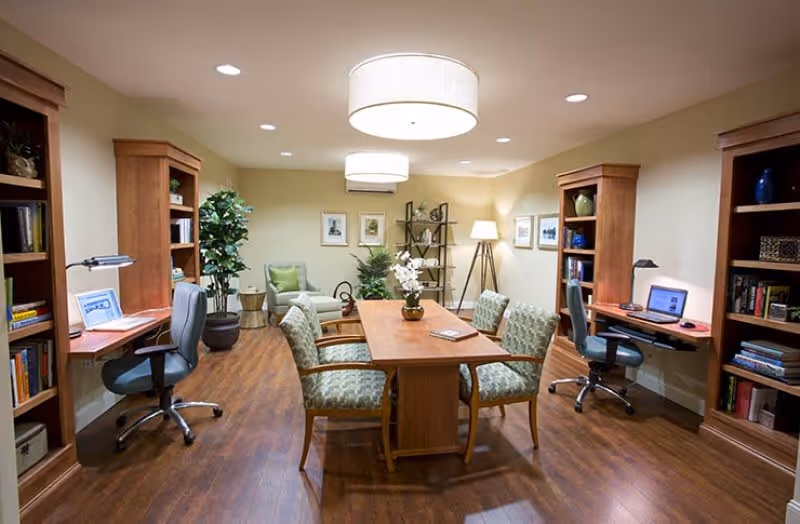 A well-lit room with wooden flooring and beige walls featuring a central wooden table surrounded by six patterned chairs. On both sides of the room, there are built-in wooden bookshelves with desks and office chairs, each desk equipped with a laptop and desk lamp. The room is decorated with potted plants, framed pictures on the walls, and a standing lamp in the corner.