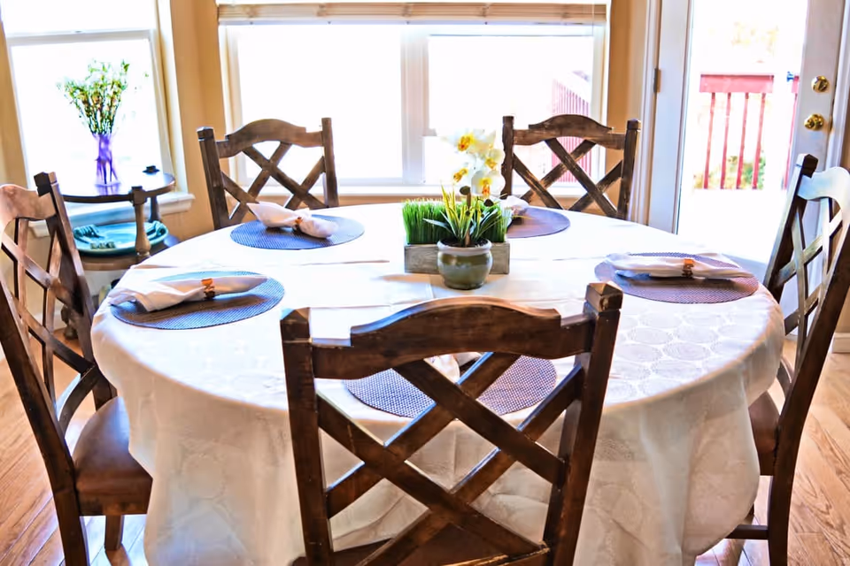 Round dining table set with six wooden chairs, placemats, napkins, and a floral centerpiece in a bright dining room.